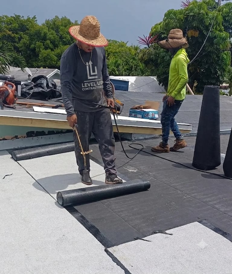 Worker in straw hat and Level Up shirt using roller tool to lay black roofing material, with assistant in yellow-green vest nearby on commercial roof