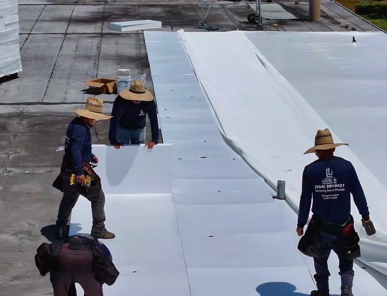 Workers installing white roofing material on a flat roof, wearing hard hats and safety gear, view from above